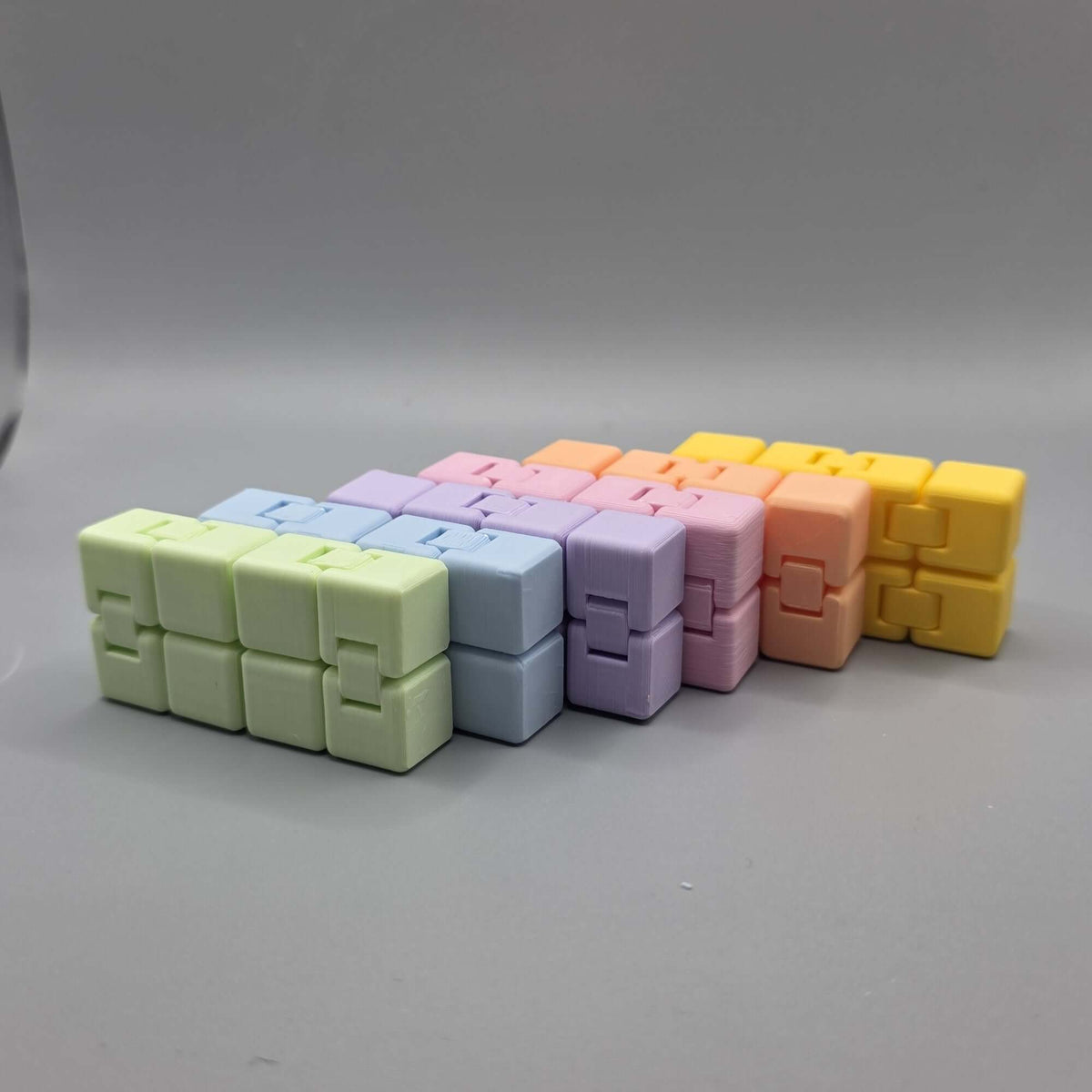 A set of six small cube-shaped puzzle toys arranged in a row on a smooth gray surface. The colors progress from light green, pale blue, lavender, pink, peach, to bright yellow.