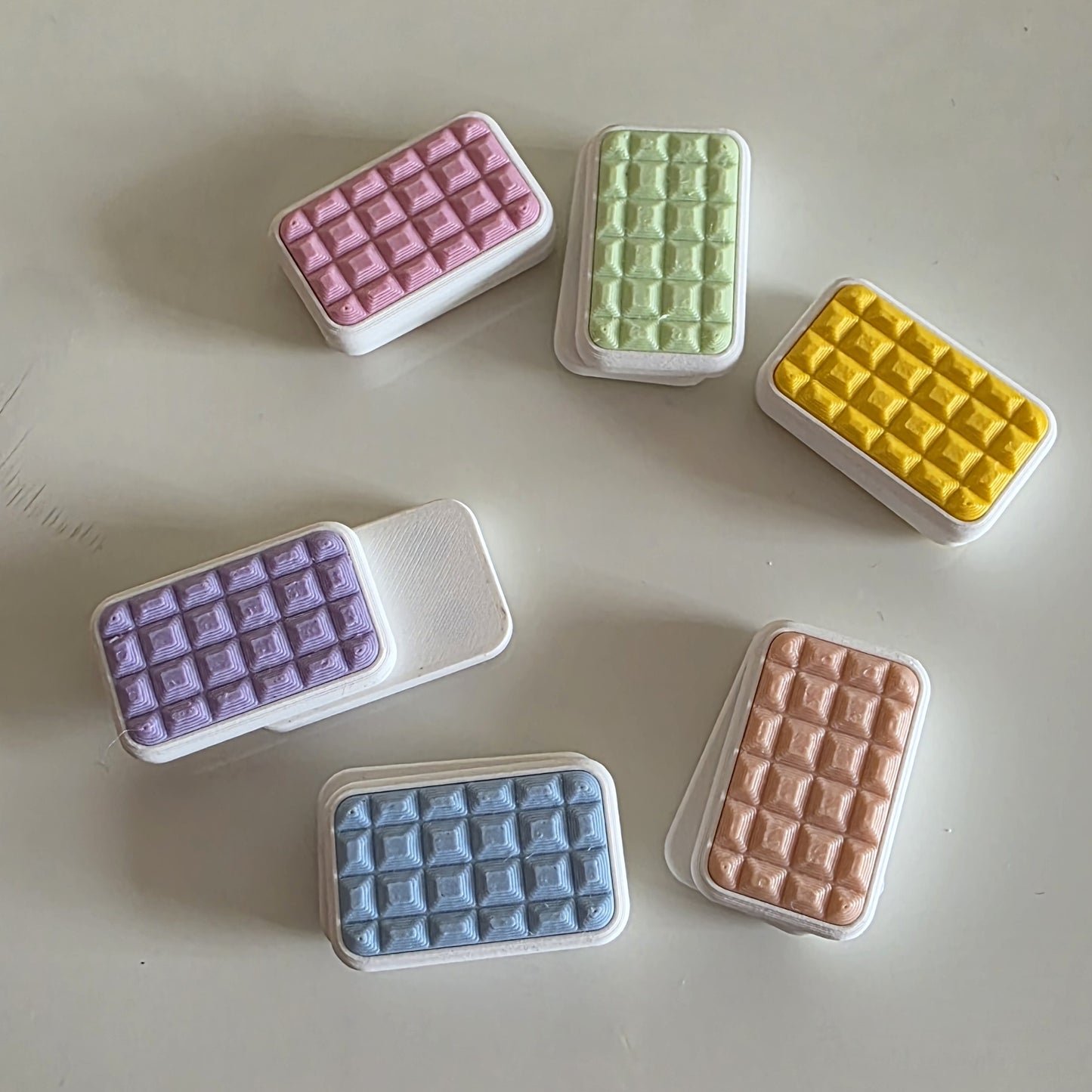 Six rectangular wax melts or soap bars in assorted pastel and bright colors, arranged on a light beige surface. The colors include pink, light green, yellow, lavender, light blue, and peach, each showing a textured grid pattern on the top half with a solid white textured side.