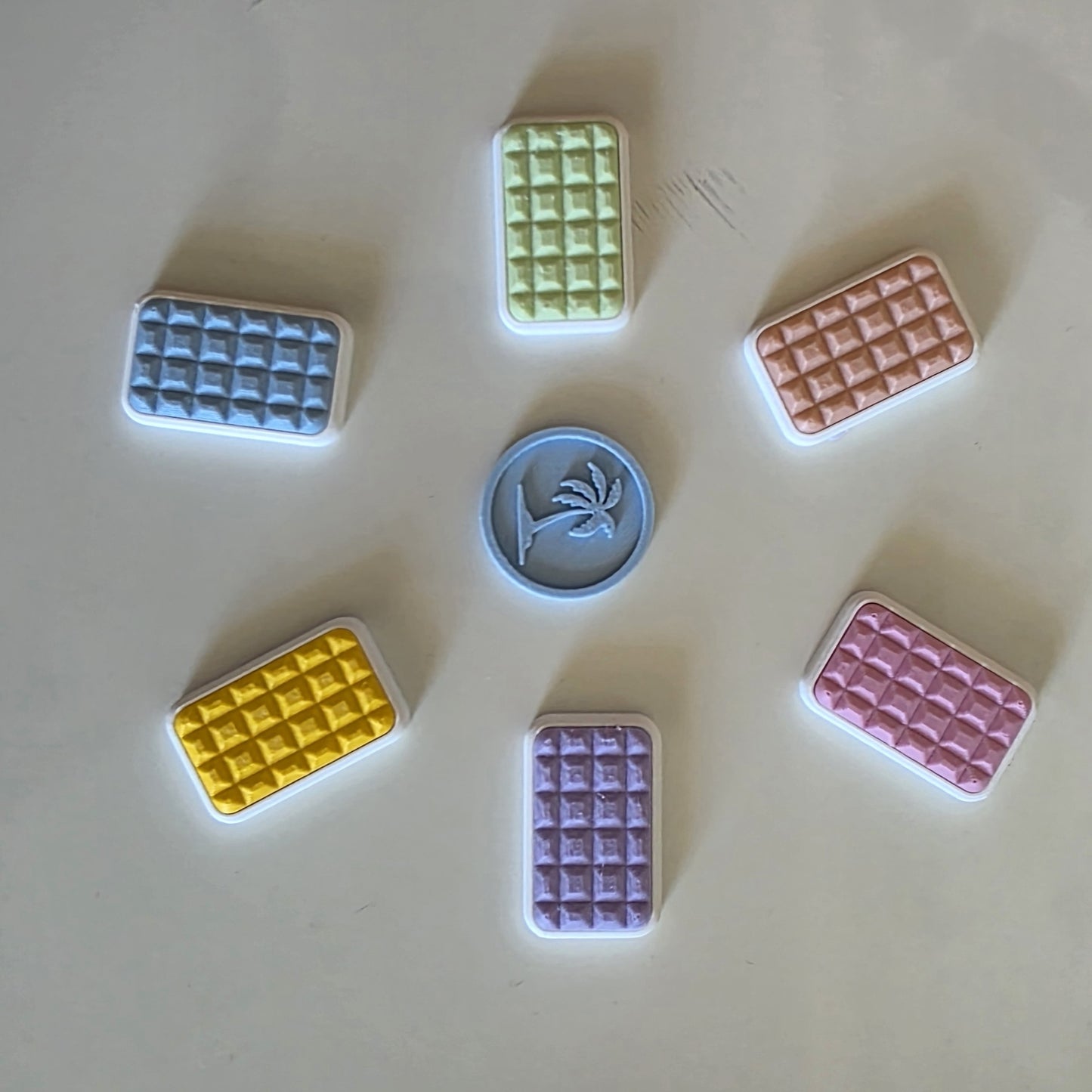 Six small rectangular waffle- or grid-patterned pieces in pastel blue, green, peach, yellow, purple, and pink are arranged around a central round blue piece with a palm tree design, on a light beige surface.