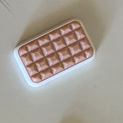 A small rectangular cosmetic item with a white border and a textured, quilted rose-gold or copper-toned top, photographed on a plain light beige surface.
