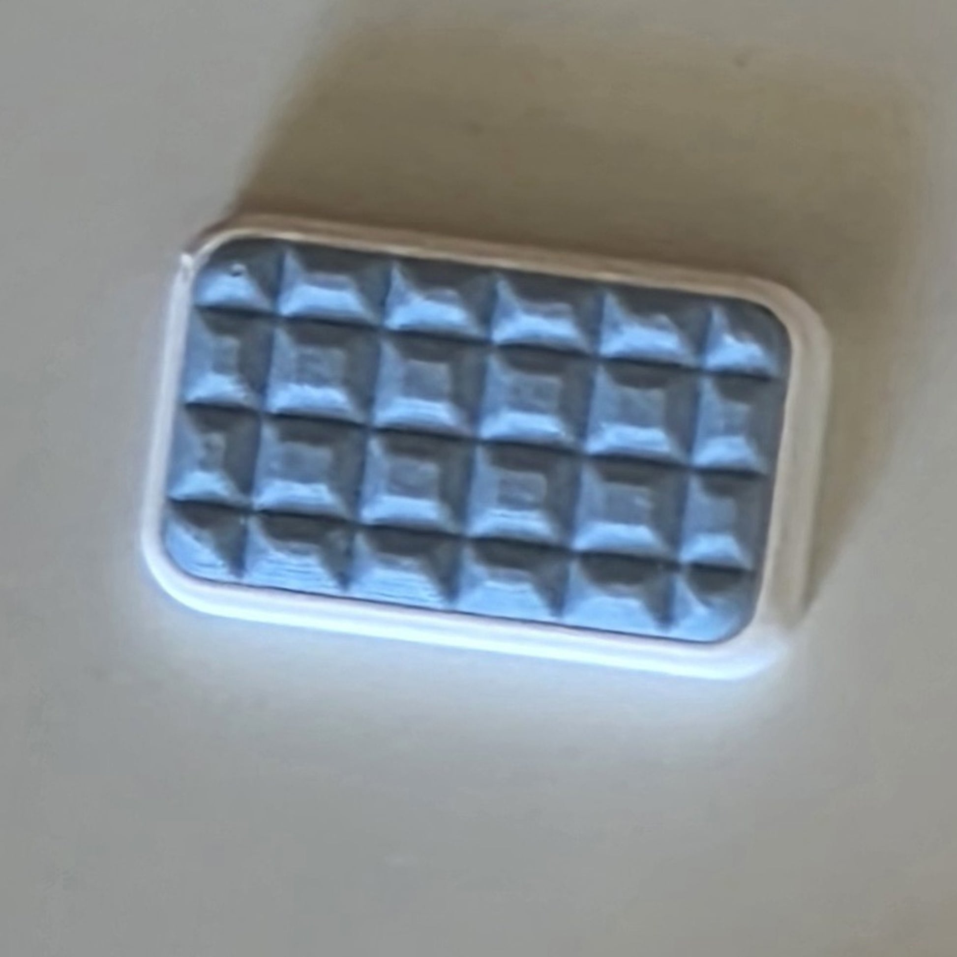 A small rectangular cosmetic item with a white border and a textured, quilted sky blue, photographed on a plain light beige surface.