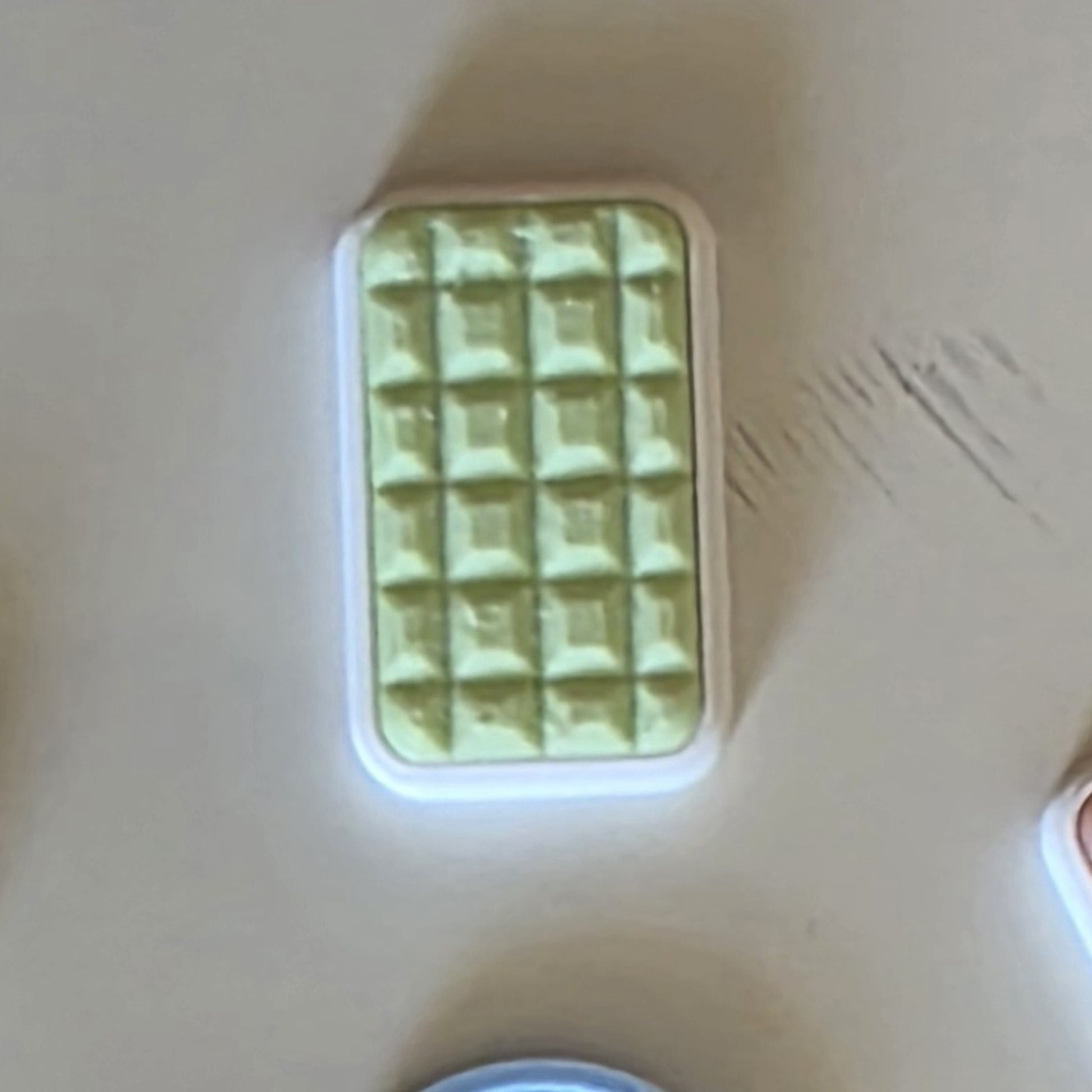 A small rectangular cosmetic item with a white border and a textured, quilted rose-gold or copper-toned top, photographed on a plain light beige surface.