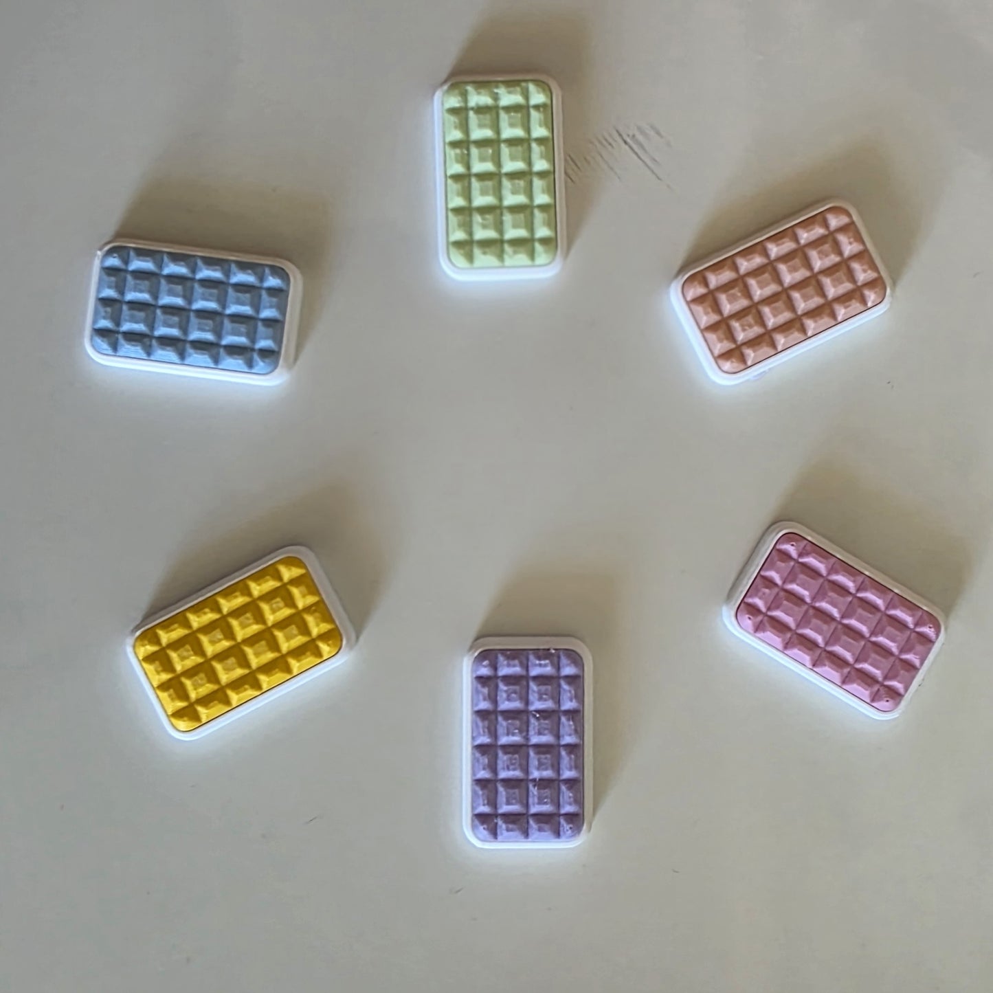 Five small rectangular items with a raised grid texture, shown in pastel blue, green, peach, yellow, and lavender on a plain light background.