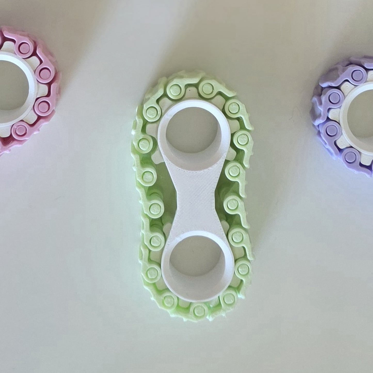 A 3D-printed circular ring tool or mold in light green and white, shown from above on a plain white surface. Partially visible at the left and right edges are similar rings in pink and purple.
