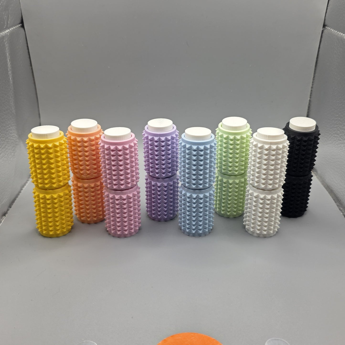 Nine textured cylindrical containers in assorted colors, including yellow, orange, pink, purple, blue, green, white, and black, arranged in two rows on a gray surface.