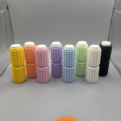 Nine textured cylindrical containers in assorted colors, including yellow, orange, pink, purple, blue, green, white, and black, arranged in two rows on a gray surface.