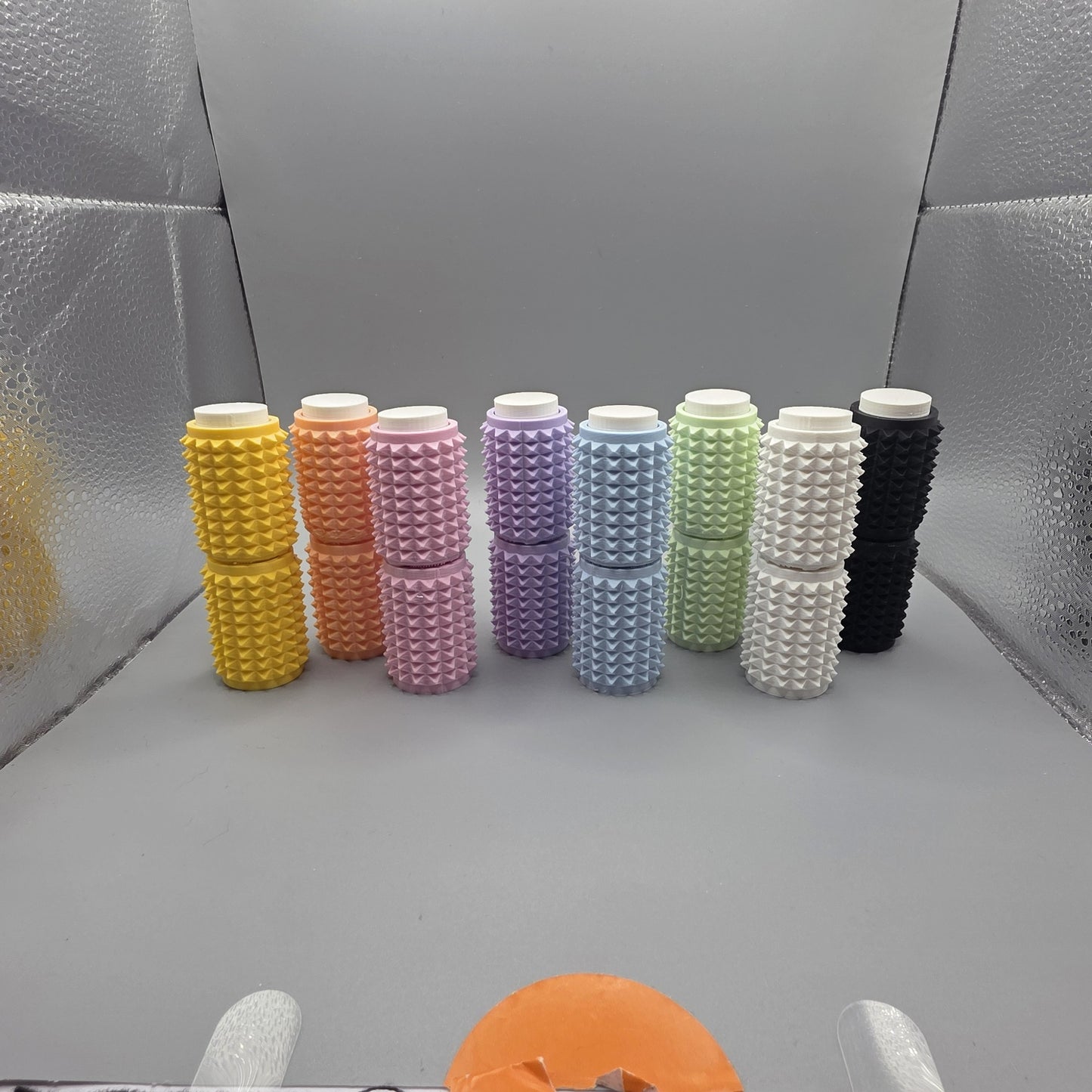 Eight textured cylindrical containers in assorted pastel and solid colors, arranged in a row on a gray surface with a reflective silver backdrop.