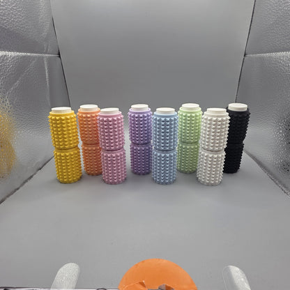 Eight textured cylindrical containers in assorted pastel and solid colors, arranged in a row on a gray surface with a reflective silver backdrop.