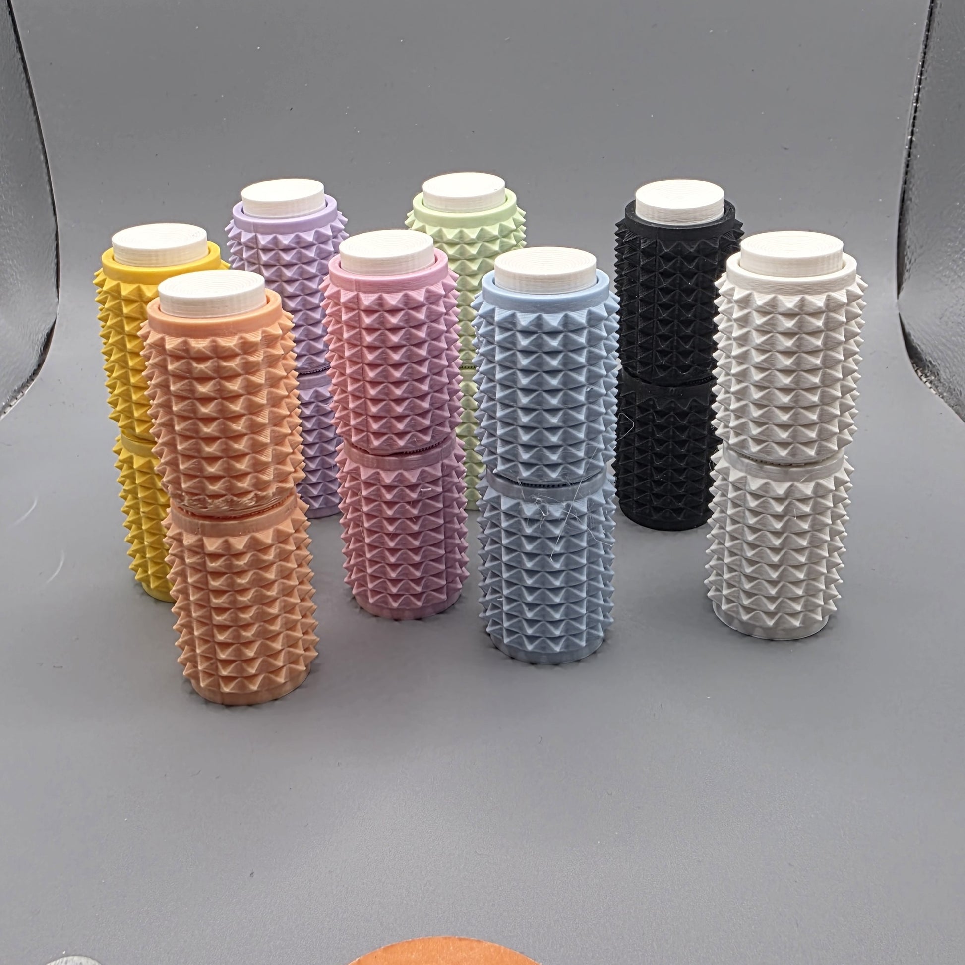 A set of eight textured cylindrical containers with white screw-on caps, arranged in two rows on a gray background. The containers come in multiple colors, including yellow, peach, pink, lavender, mint green, light blue, black, and white, each featuring a raised diamond pattern on their sides.