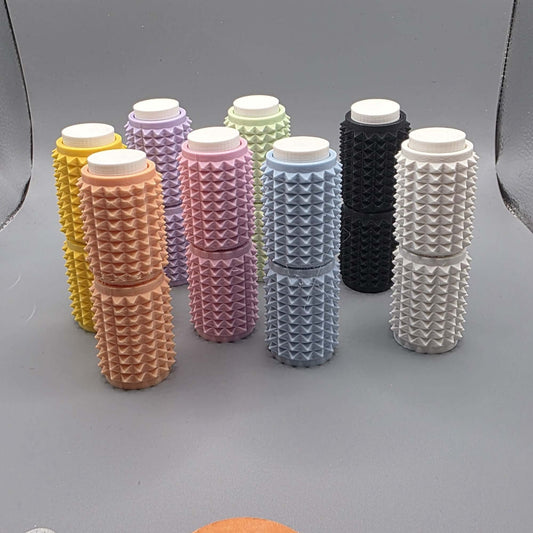A set of eight textured cylindrical containers with white screw-on caps, arranged in two rows on a gray background. The containers come in multiple colors, including yellow, peach, pink, lavender, mint green, light blue, black, and white, each featuring a raised diamond pattern on their sides.