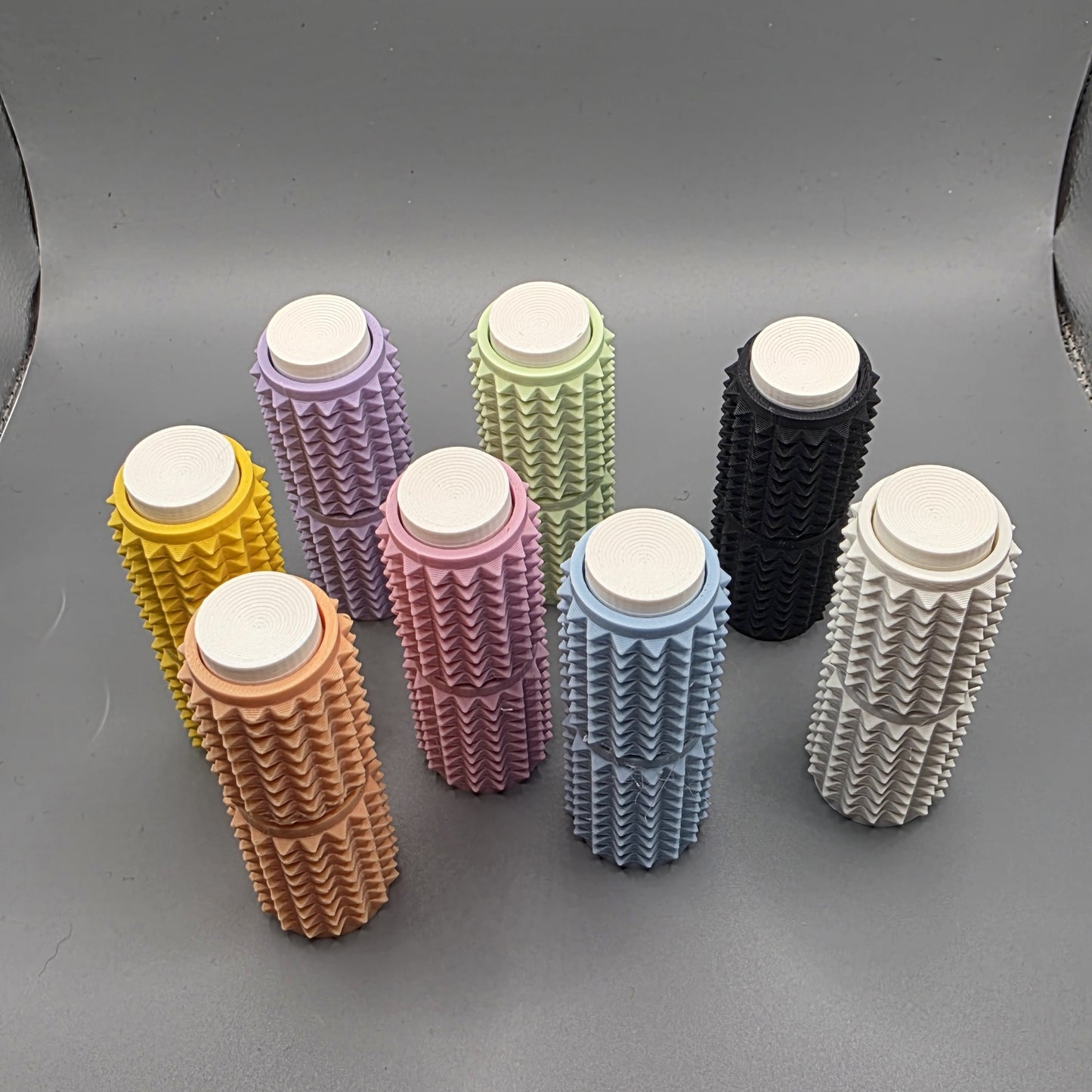 Seven textured cylindrical foam roller massage guns in assorted colors including yellow, peach, pink, purple, green, black, blue, and white, arranged on a gray background.