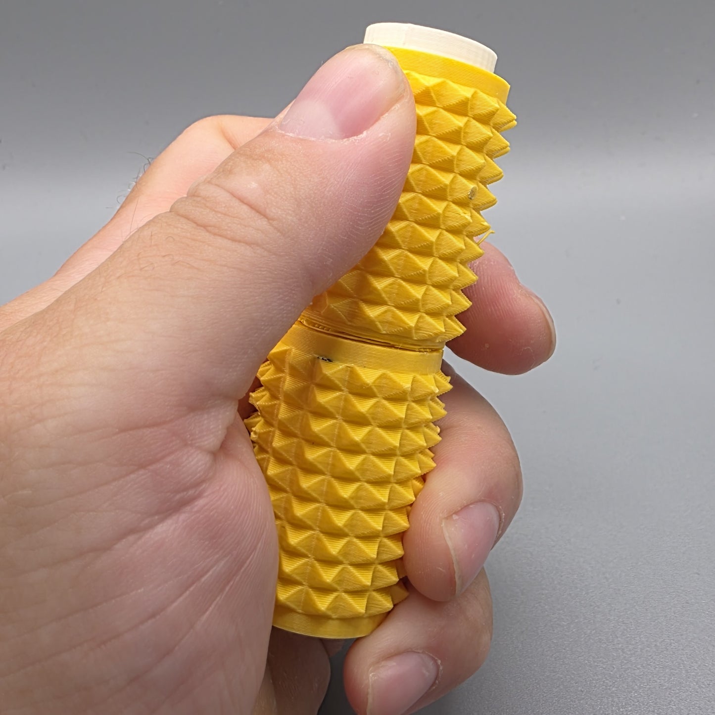 A hand holding a yellow textured cylindrical product with a ridged, spiked exterior and a white cap visible at the top, photographed against a plain gray background.