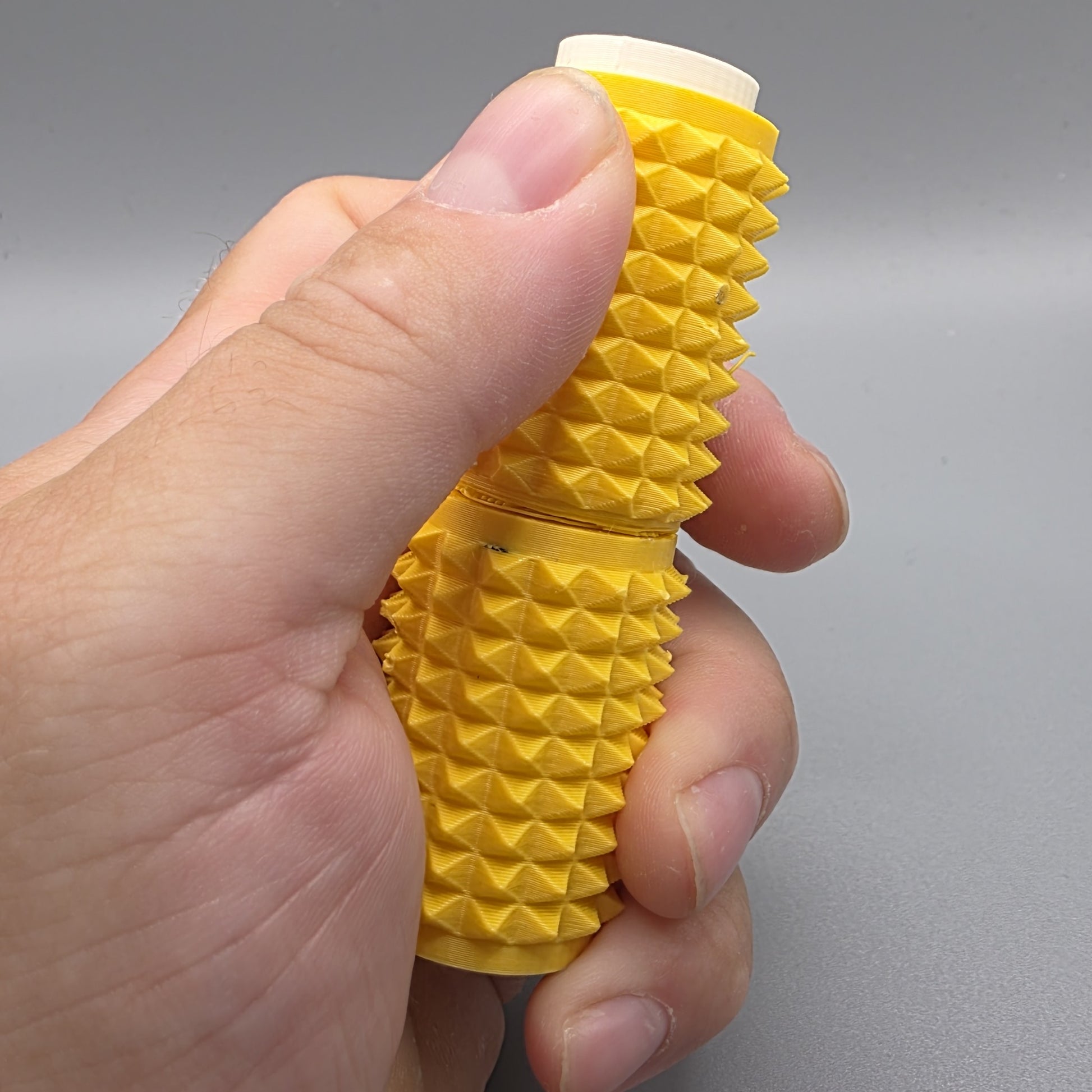 A hand holding a yellow textured cylindrical product with a ridged, spiked exterior and a white cap visible at the top, photographed against a plain gray background.
