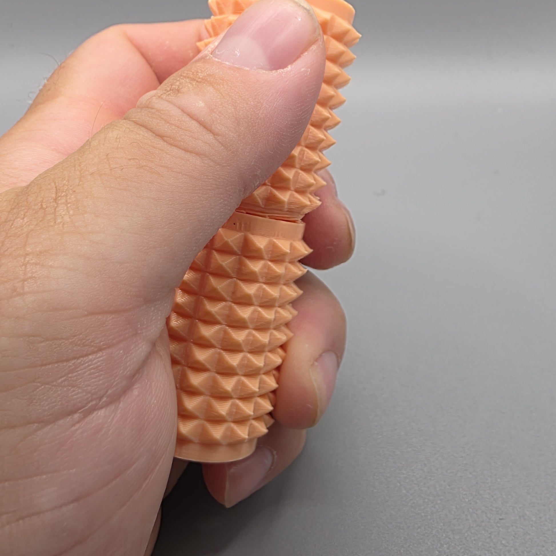 A close-up of a hand holding a textured, peach-colored silicone or rubber object with a ridged, spiky surface against a plain gray background.