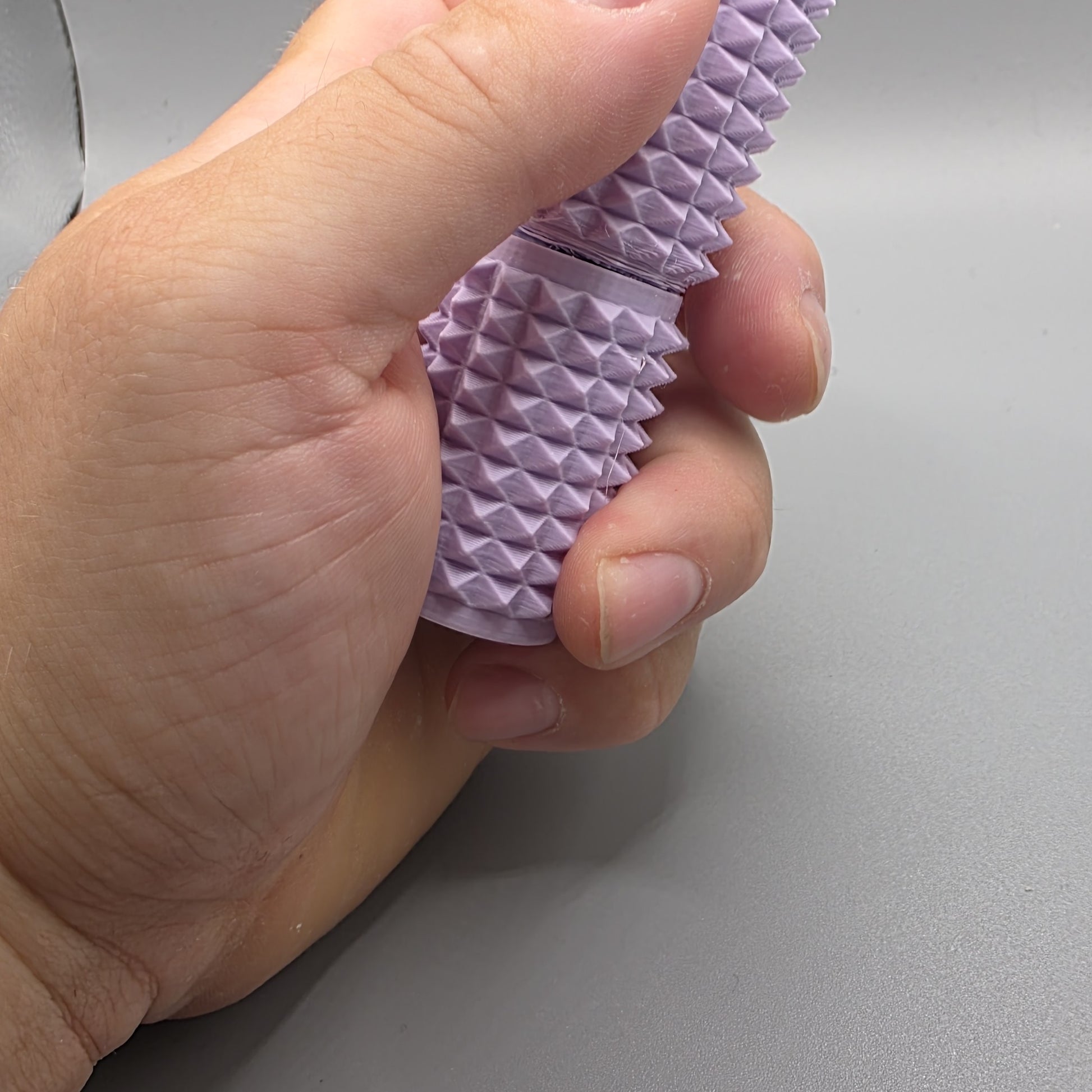 A close-up of a hand holding a lavender purple textured silicone sleeve with a pyramid pattern, set against a plain gray background.