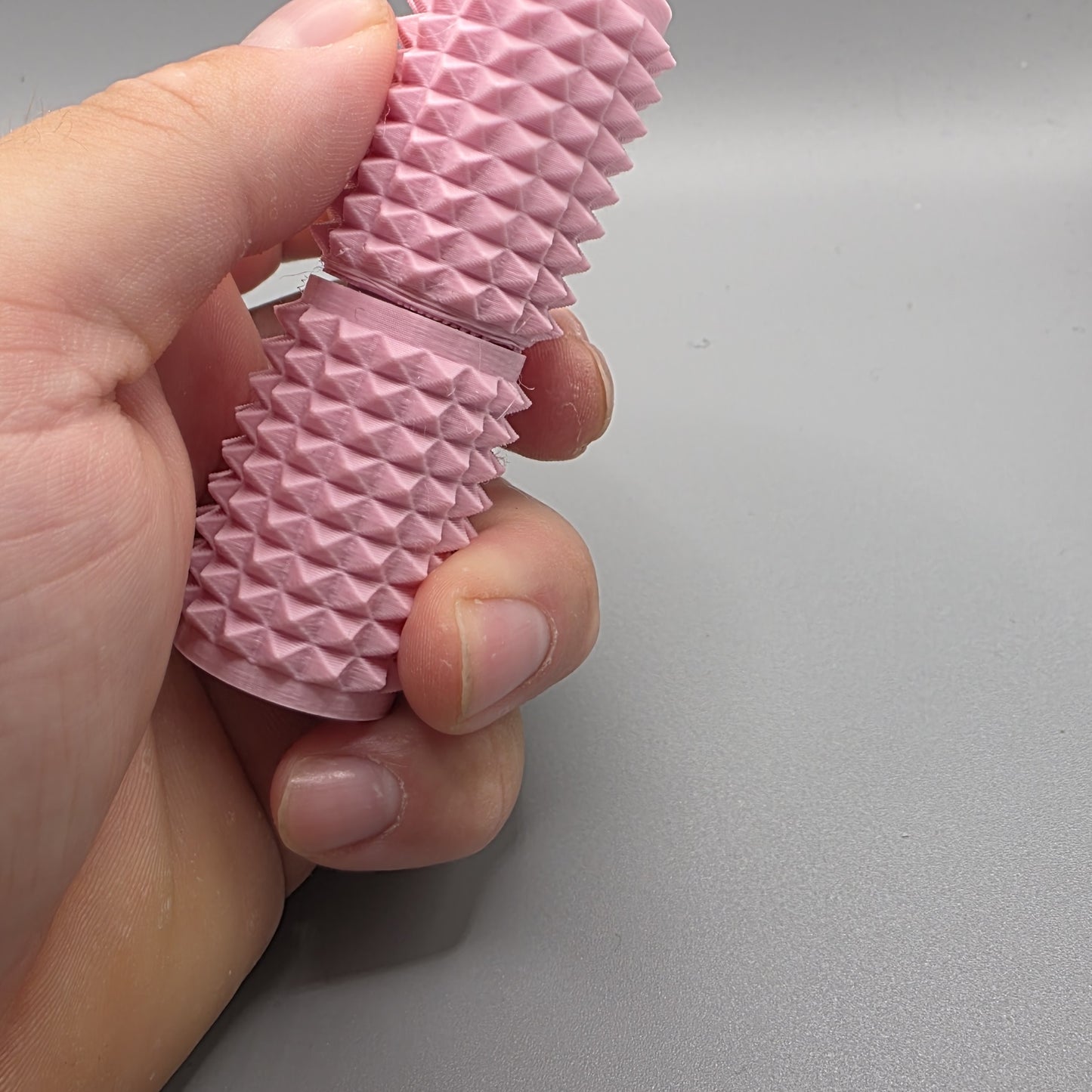A close-up of a hand holding a pink textured silicone sleeve with a ridged, pyramid-patterned surface against a plain gray background.