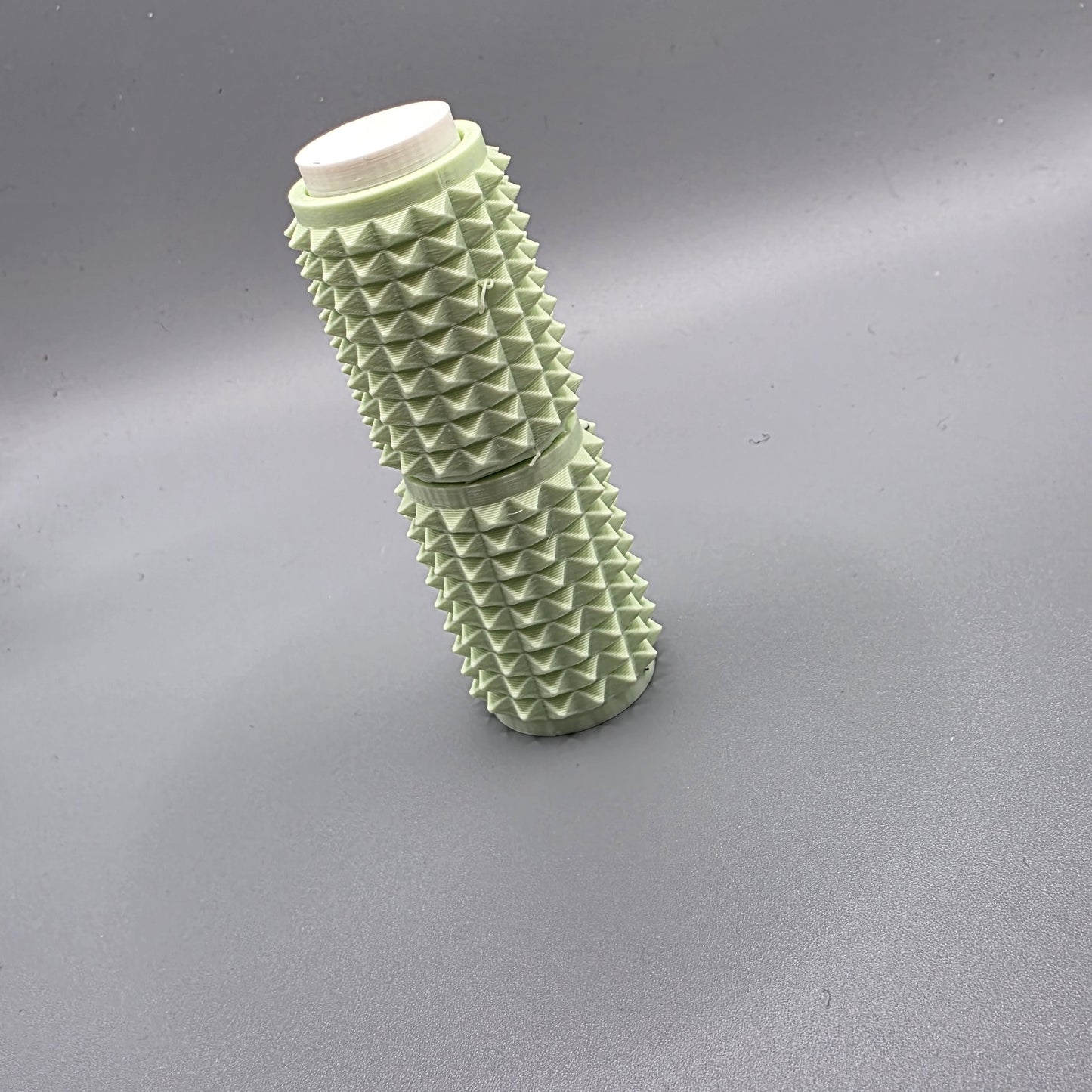 A light sage green textured cylindrical container with a white screw-on cap, standing upright on a neutral gray background.