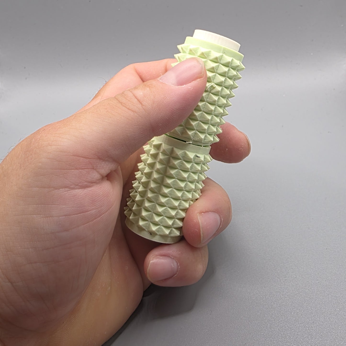 A hand holding a light green textured cylindrical device with a ridged, spiky exterior and a white inner core, shown against a neutral gray background.