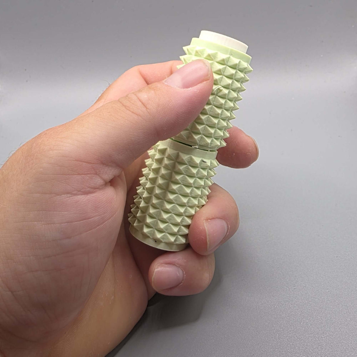 A hand holding a light green textured cylindrical device with a ridged, spiky exterior and a white inner core, shown against a neutral gray background.