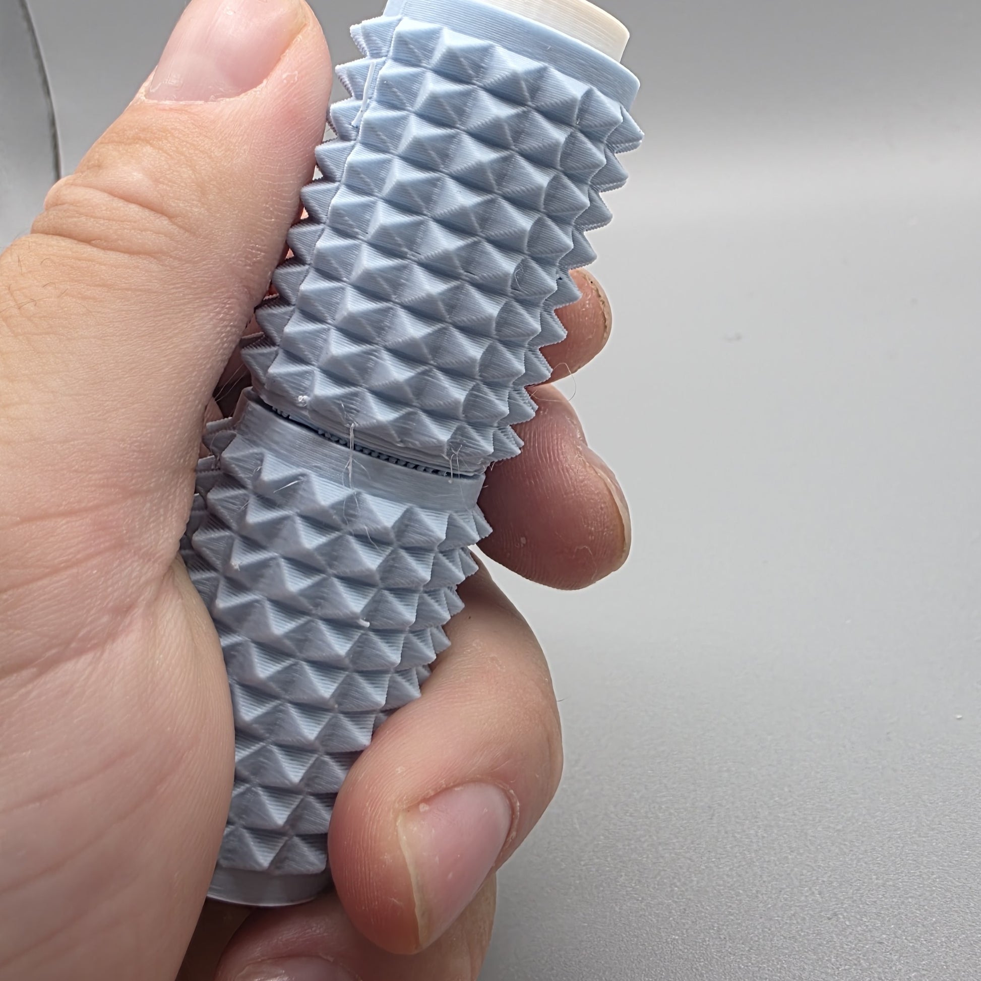 A close-up of a hand holding a textured, ribbed cylindrical cosmetic tube, likely a lip balm or cream stick, against a plain light gray background.
