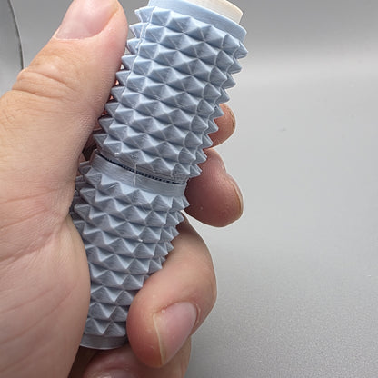 A close-up of a hand holding a textured, ribbed cylindrical cosmetic tube, likely a lip balm or cream stick, against a plain light gray background.