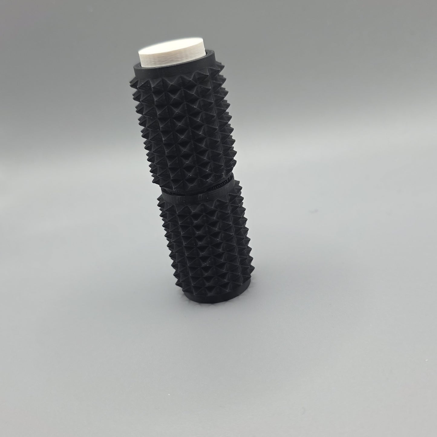 A black textured cylindrical object with a white circular top, standing upright on a smooth gray surface.