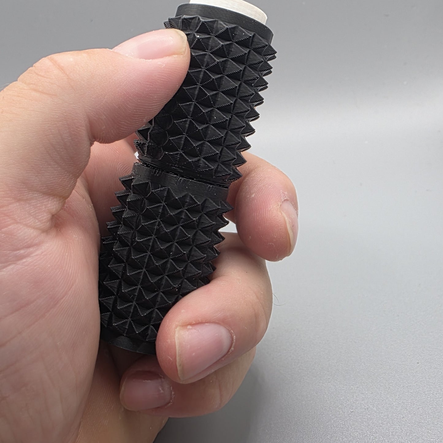 A close-up of a hand holding a black textured cylindrical product with a ridged, pyramid-like surface and a small white tip visible at the top, against a plain light gray background.