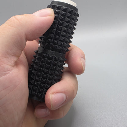 A close-up of a hand holding a black textured cylindrical product with a ridged, pyramid-like surface and a small white tip visible at the top, against a plain light gray background.