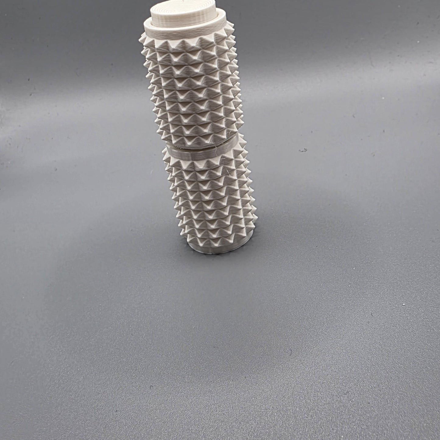 A white textured cylindrical container with a ribbed, spiky surface and a screw-on cap, standing upright on a gray surface.