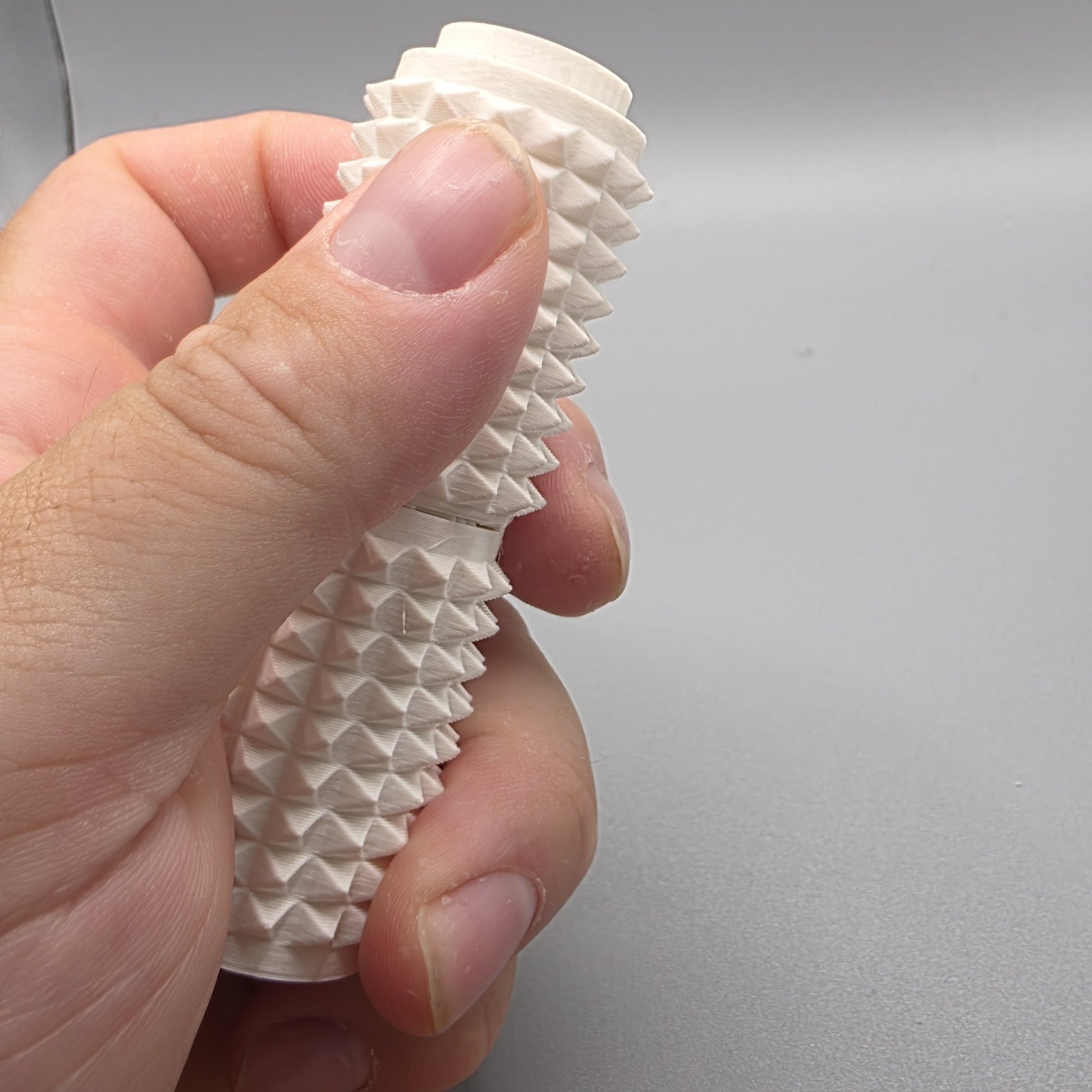 A close-up of a hand holding a textured white cylindrical object with a ridged, spiky surface against a plain gray background.
