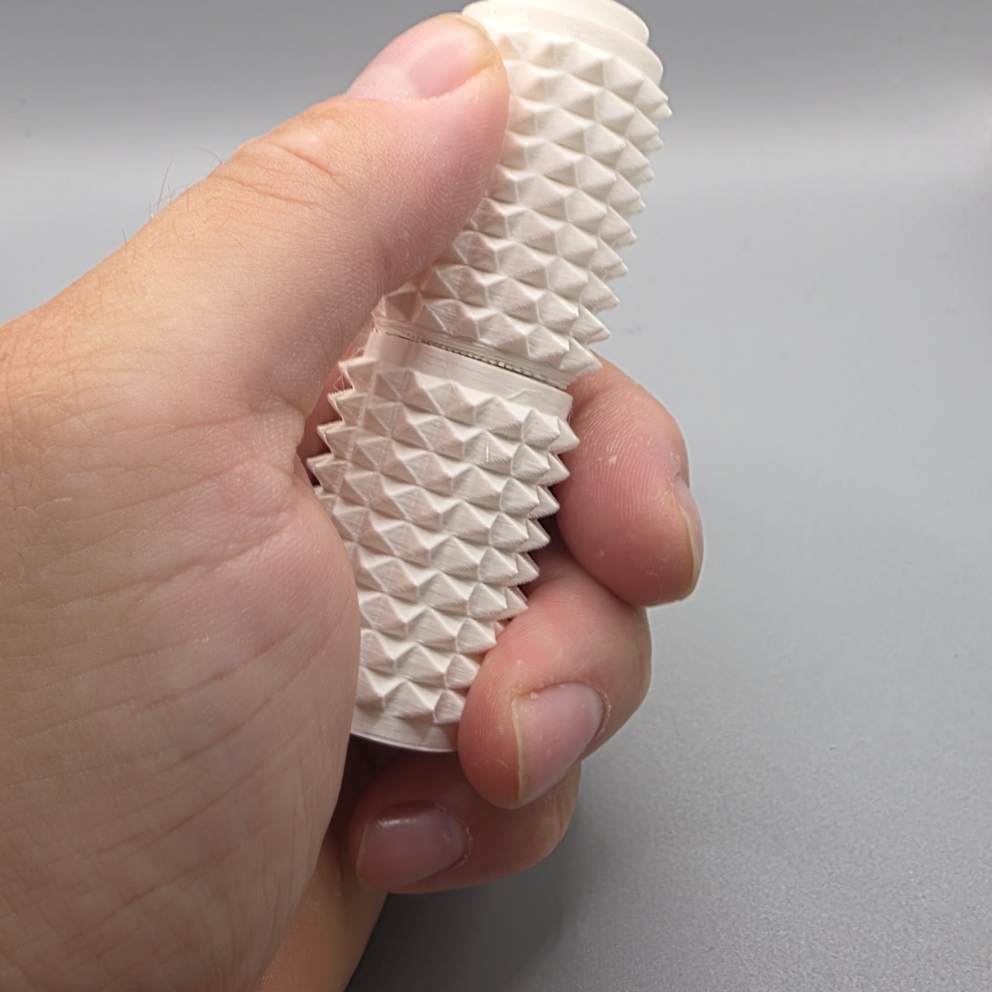 A close-up of a hand holding a textured, off-white silicone sleeve with a ridged, pyramid-like pattern against a neutral gray background.