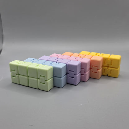 A set of six small cube-shaped puzzle toys arranged in a row on a smooth gray surface. The colors progress from light green, pale blue, lavender, pink, peach, to bright yellow.