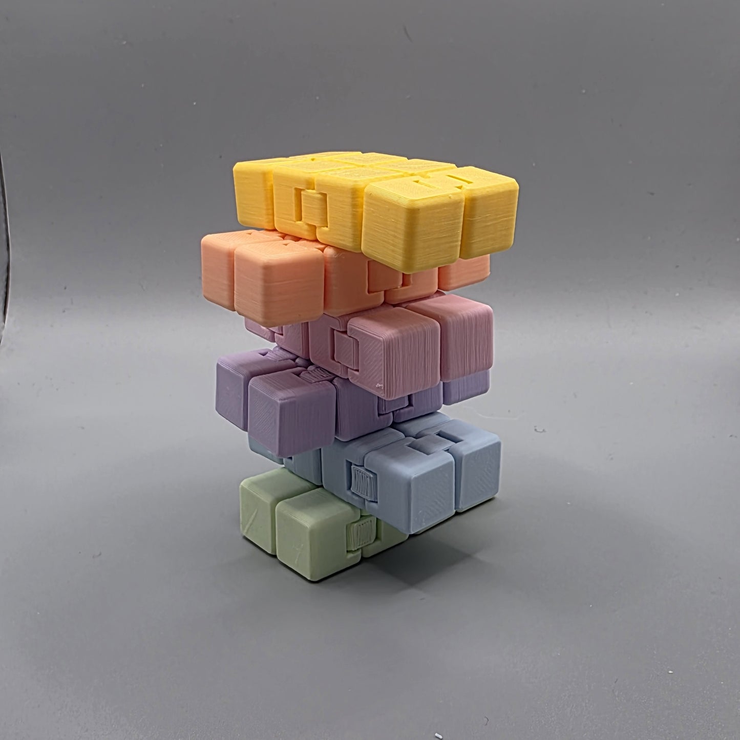A stack of modular 3D-printed cube blocks in pastel colors, including yellow, peach, pink, lavender, light blue, and sage green, arranged on a neutral gray background.