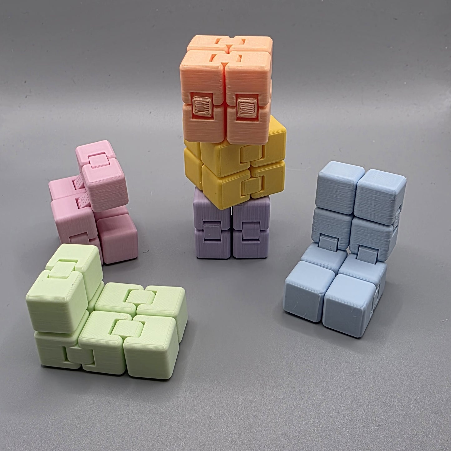 Four 3D-printed cube-based toy structures in pastel colors, including light green, pink, orange/yellow, and light blue, arranged on a neutral gray background.